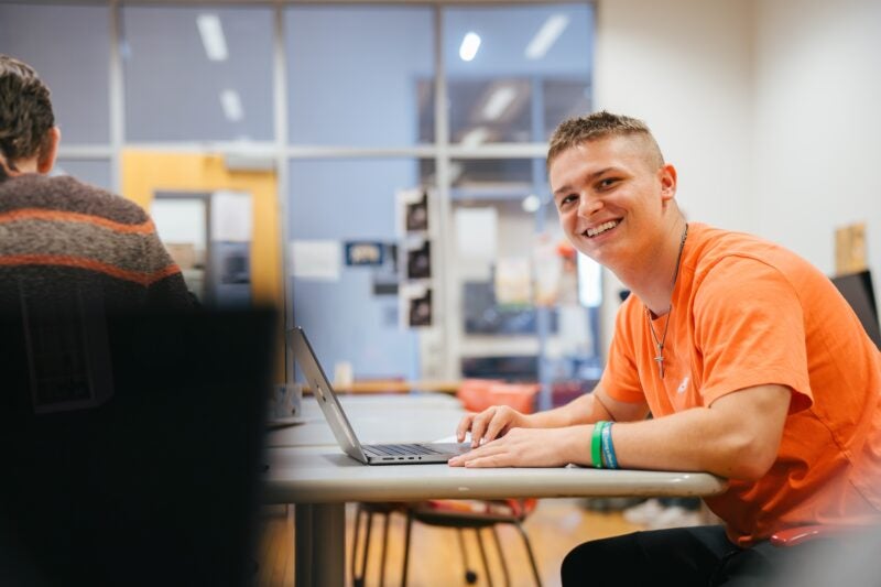 student working on a laptop in a classroom and smiling at the camera