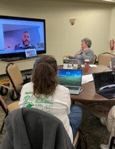 a group of students and educators interacting with a livestream on a projector while sitting at a table at Marshall University