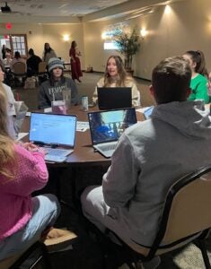 a group of individuals including students and educators conversing and presenting while sitting at a table at Marshall University
