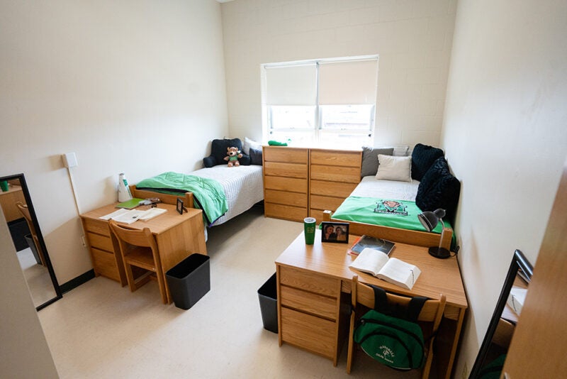 A sample residence hall room at Marshall University includes furniture setup for two students with green and black, Marshall themed bedding.