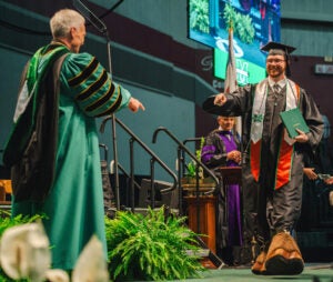 Gauge Goff walks across the stage at Marshall University's Spring Commencement wearing Marco's hooves. President Brad D. Smith points and smiles as he recognize's the hooves.