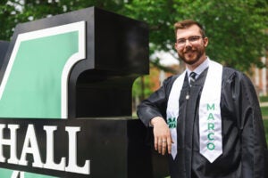 Gauge Goff wears his graduation gown and Marco stole while smiling next to the Block M sign outside of Marshall University's Old Main.