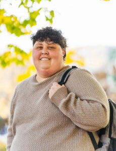Marshall student, Mika Sheppard smiles as she holds the strap of her backpack while standing outside in front of fall trees
