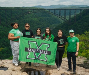 Students wearing Marshall University attire hold a Marshall flag and smile for a photo in front of the New River Gorge Bridge