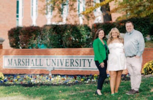 Haley Prather with her dad and step-mom in front of a brick Marshall University sign