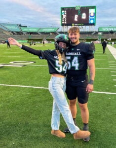 Allie Ingles wears a football helmet and stands next to a Marshall player on the Marshall University football field