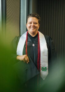 Alexie Adkins smiles while holding a graduation cap and wearing a graduation gown and Marshall University stole
