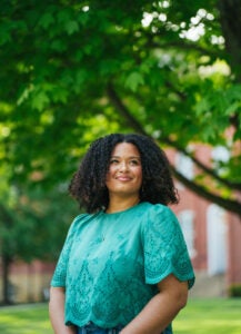 Haleigh Muncy wears a green shirt and stands under a tree looking up with a red brick building in the background