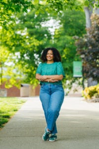 Haleigh Muncy wears a green shirt and stands on a sidewalk with her arms crossed. A green Marshall University banner hangs from a light stand in the background