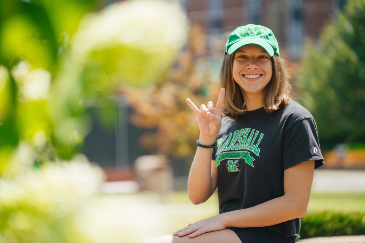 student outside on campus smiling at the camera and showing a Go Herd sign with her hands