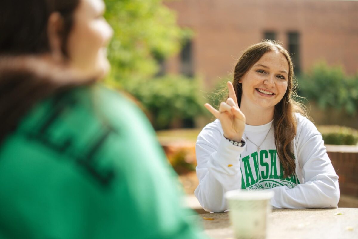 student smiling outside the Memorial Student Center showing a Go Herd symbol with her hands