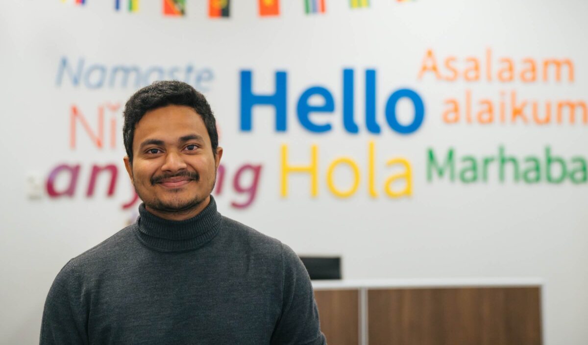 student in East Hall smiling at the camera with Hello in different languages on the wall behind
