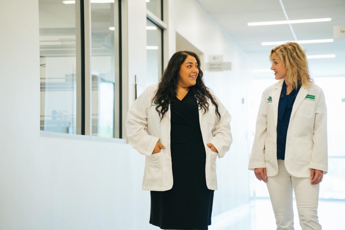two students in lab coats walking down a hallway talkiing