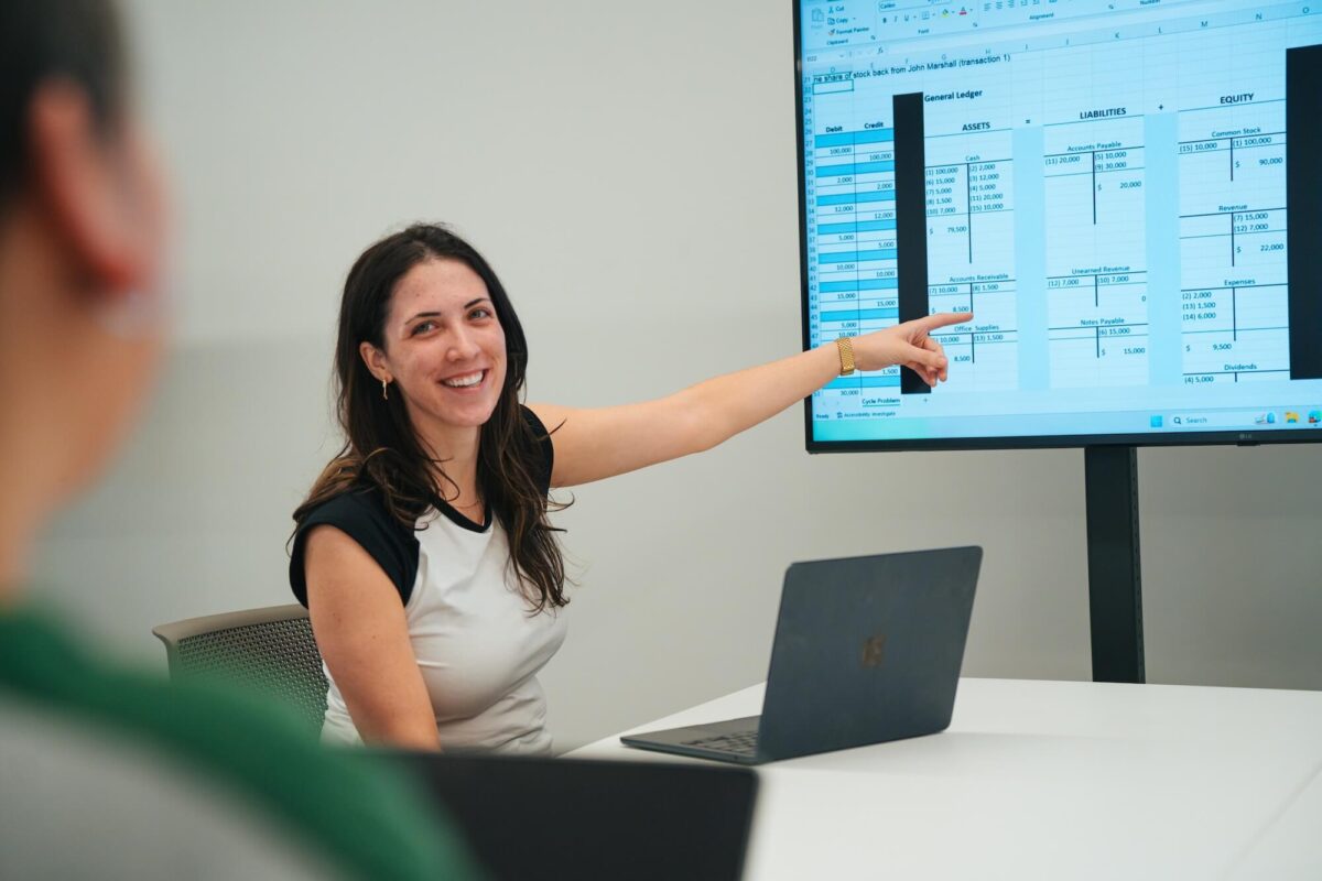 student working on a laptop pointing to a spreadsheet on a large screen