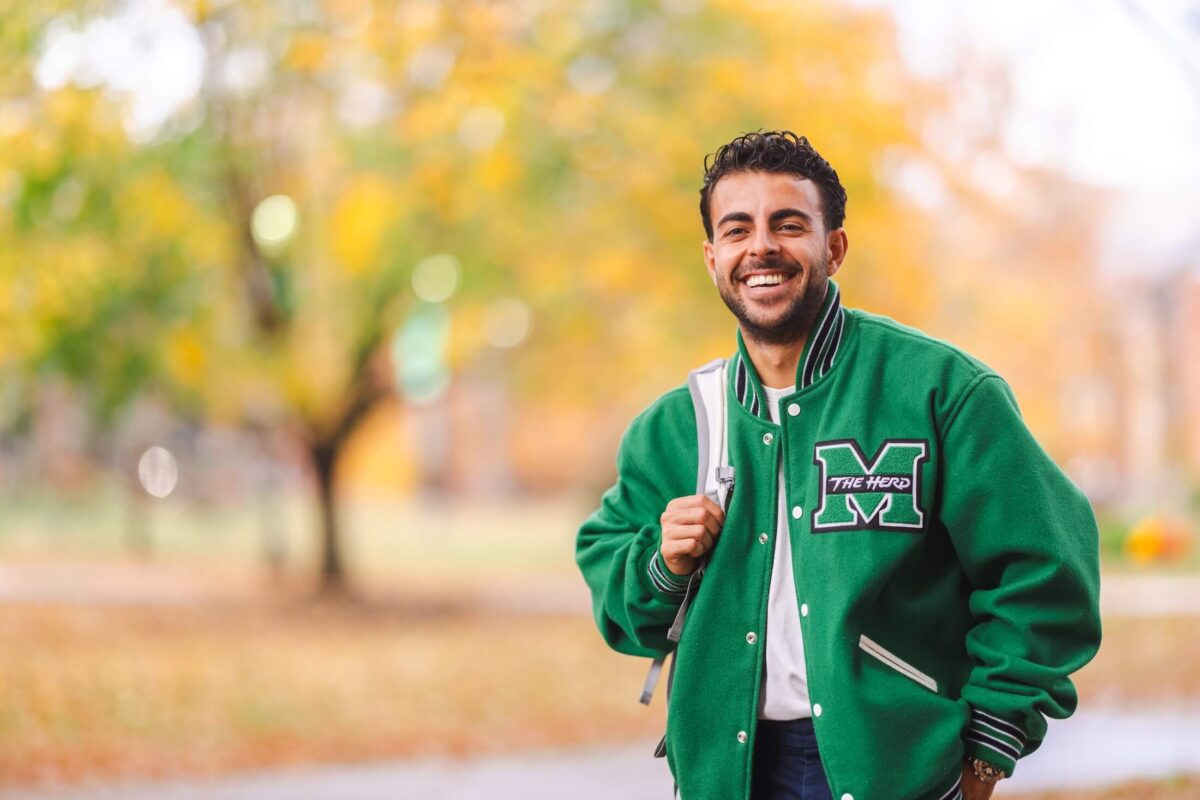 student in a Marshall letterman jacket smiling outside on campus with a backpack on