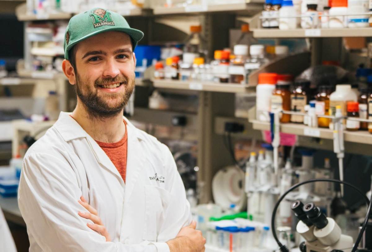 student in a labcoat in a lab smiling at the camera