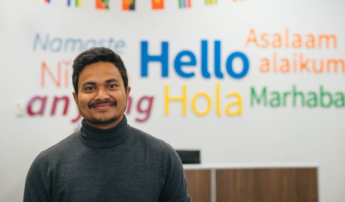 student in East Hall smiling at the camera with Hello in different languages on the wall behind