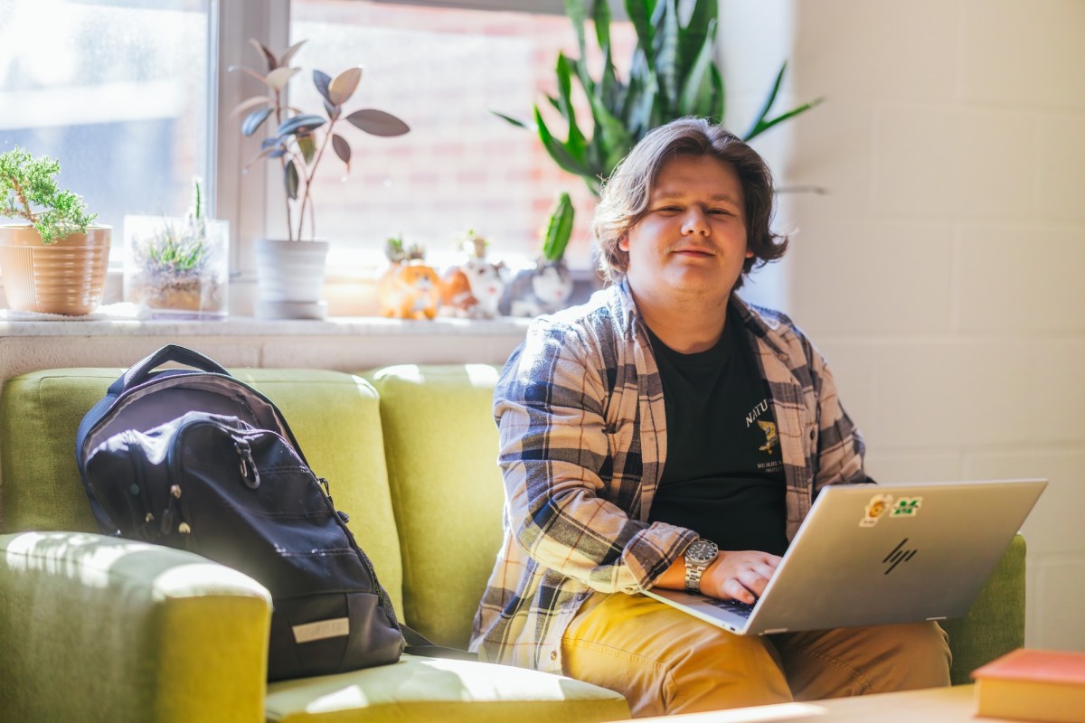 student working on a laptop on a couch in front of a window in a residence hall