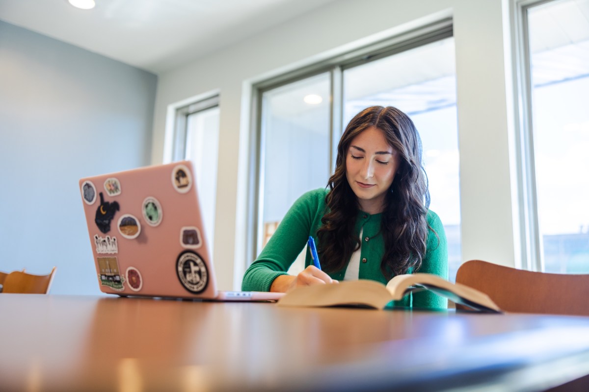 student working in a study room reading a book and working on a laptop