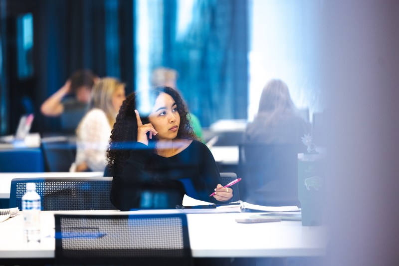 A student sits at a table in a modern classroom holding a pen and looking thoughtfully toward the front of the room during class. Open notebooks and a laptop sit on nearby tables, while other students are blurred in the background.