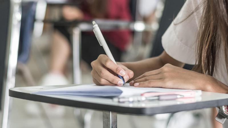 A young girl sits at a desk completing a written test or assignment. Image shows a closeup view of the student writing.
