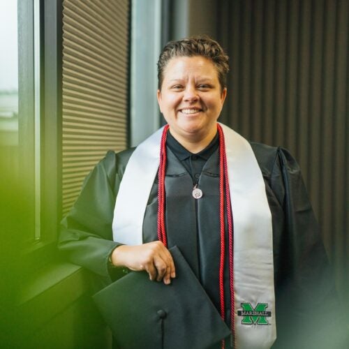 Alexie Adkins stands next to a window smiling while wearing her graduation gown, cords and stole. She is holding her graduation cap.