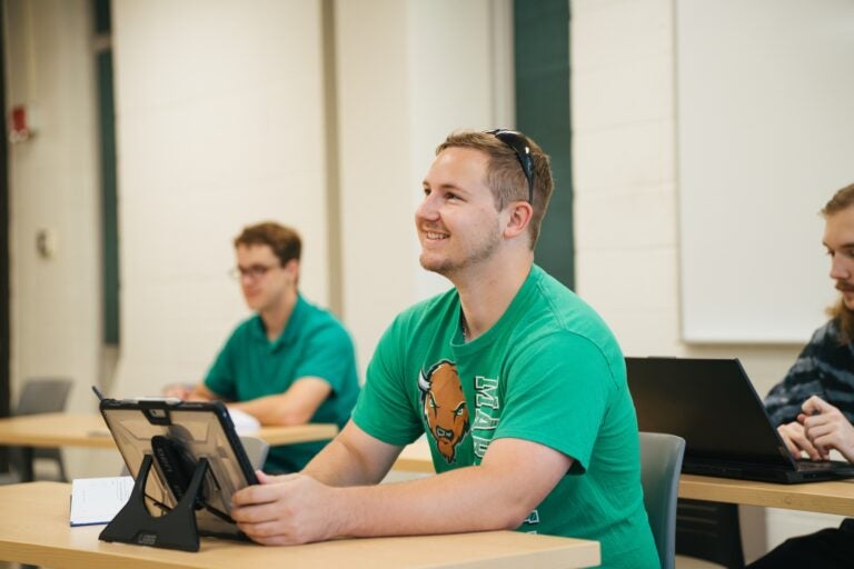 student sitting in a class on an iPad and looking toward the front of the class