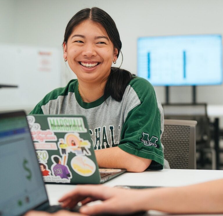 student using a laptop in a classroom smiling and looking at another student