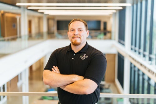Dylan Glasscock wears a black polo shirt with Greek letters while standing with his arms crossed in a bright, modern building.