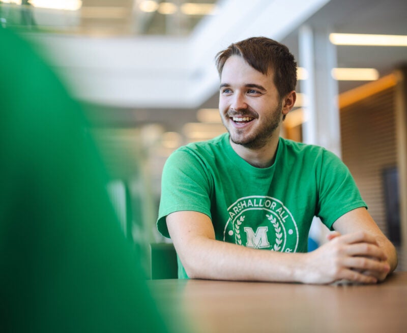 A student wearing a green Marshall shirt sits at a table smiling at someone else