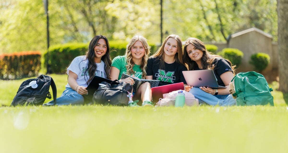 Students wearing Marshall University attire sit together in the grass with their laptops and notebooks.
