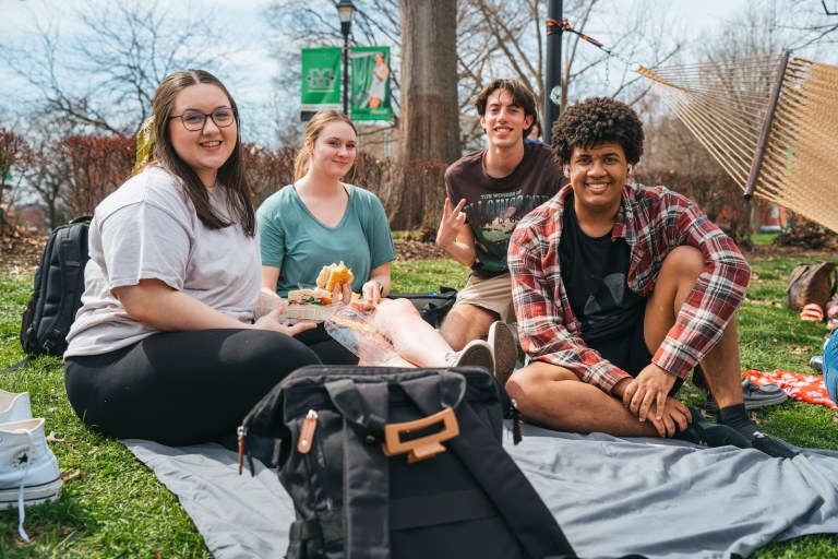 group of four students on Buskirk Field sitting on a blanket and smiling at the camera