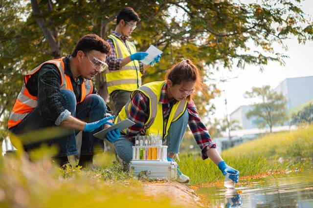 Workers wearing safety equipment collect and record water quality samples from a campus pond.