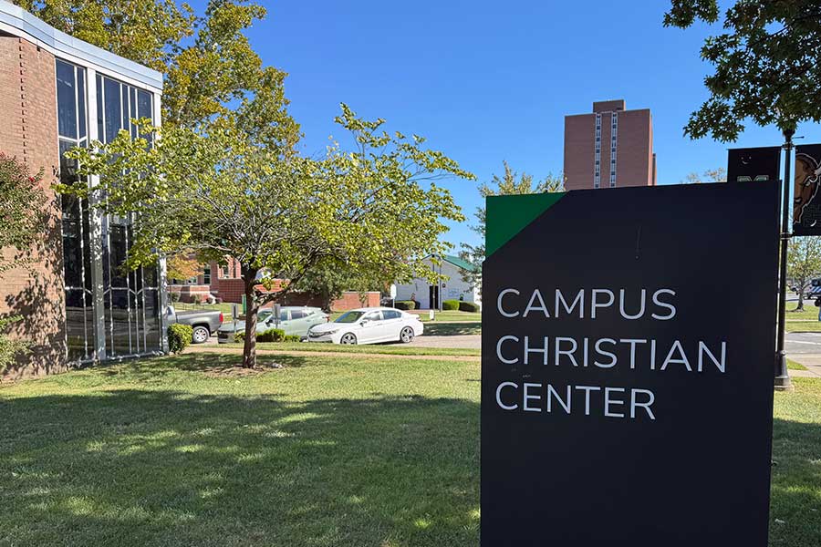 Baháʼí Martyrs Memorial Tree, an Eastern Redbud, growing on the lawn beside the Campus Christian Center, with a Campus Christian Center wayfinding sign visible in the foreground.