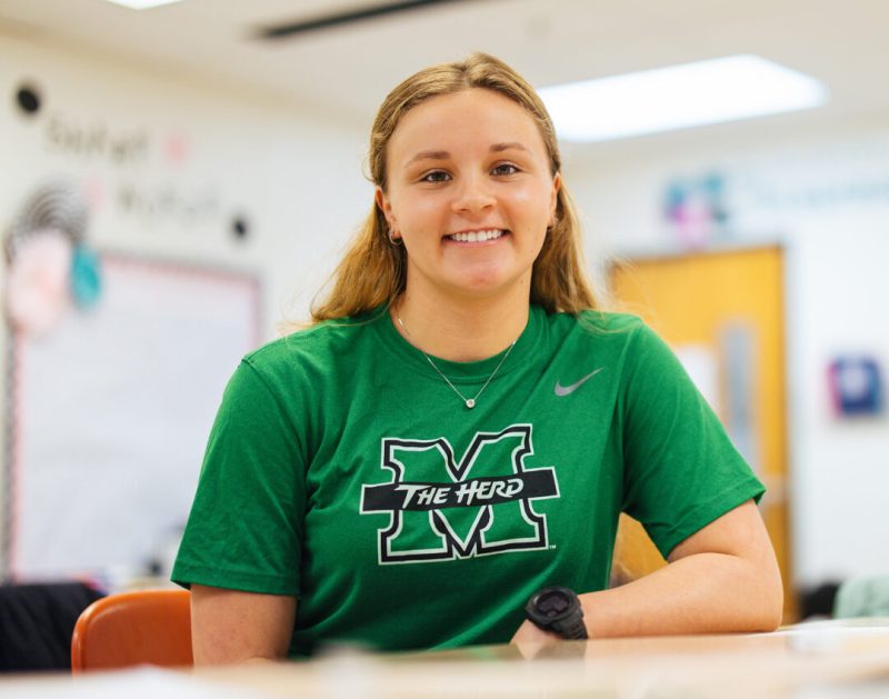 A student wearing a Marshall University t-shirt smiles while sitting at a table in a classroom