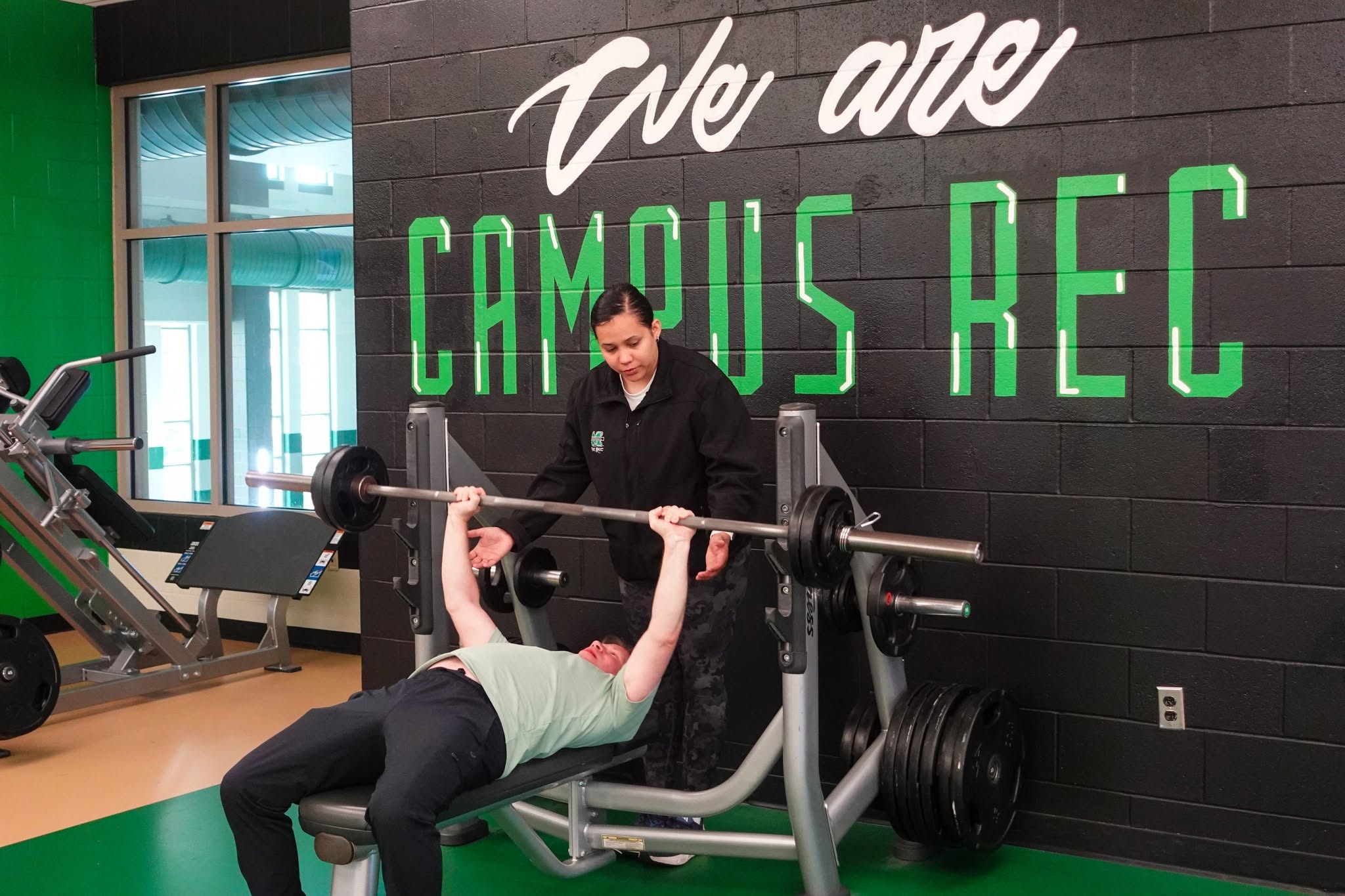 A Marshall University fitness staff member spots a student performing a bench press exercise in the university recreation center weight room.