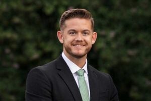 Professional headshot of Bradley Muncy wearing a dark suit, white shirt, and light green tie, standing outdoors with green foliage softly blurred in the background