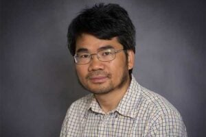 Professional headshot of Marshall University biology professor Dr. Guo-Zhang Zhu wearing glasses and a checkered shirt against a gray studio background.