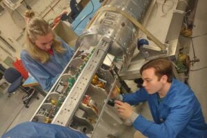 Two students in a lab environment secure soil sample containers inside an open rocket payload section, surrounded by wiring and instrumentation, during pre-launch preparations for a NASA sounding rocket