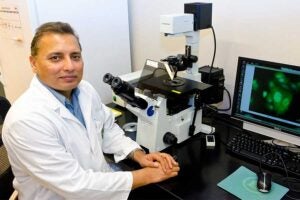 Dr. Jagan Valluri in a white lab coat seated at a desk beside a microscope, with a computer monitor displaying magnified green cell images, illustrating hands-on cancer research work