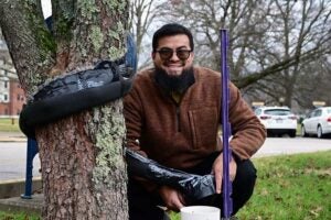 Jorge Vera crouches beside a campus tree equipped with his rainwater collection devices and tubing, holding a measuring tool near a small collection container at the base, illustrating field research on how trees capture and redirect rainwater in an urban environment