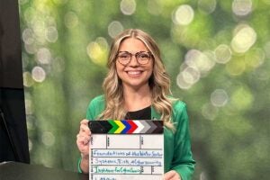 Cyber Forensics and Security graduate Lakan Sheets holds a production clapperboard while helping create video content for Marshall University's Institute for Cyber Security in a recording studio with a backdrop.