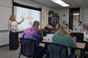 Faculty members participate in a professional development workshop, with one presenter standing beside a projected slide and speaking to colleagues seated around tables in a classroom. Laptops, notebooks, and instructional materials are visible as participants discuss STEM teaching and learning strategies.