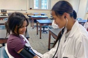 Participant wearing a stethoscope and white lab coat practices taking blood pressure on another participant during a hands-on activity in a classroom at the Marshall University College of Science Pre-Med Camp