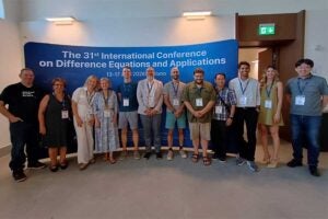 Attendees at the 31st International Conference on Difference Equations and Applications pose for a group photo in front of a conference display in Milan, Italy. Marshall University assistant professor of mathematics Dr. Tom Cuchta, wearing a green shirt and khaki shorts, stands near the center of the group.