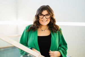 Portrait of Marshall University mathematics student Piper Cook standing beside an indoor railing, wearing a green blazer and glasses, with a bright, modern campus interior in the background