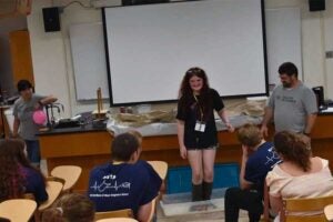 A participant stands in a clear plastic container filled with a non-Newtonian fluid during a physics demonstration while other students watch from classroom seats. Demonstration materials and instructors are visible at the front of the classroom during a Marshall University HSTA science event.