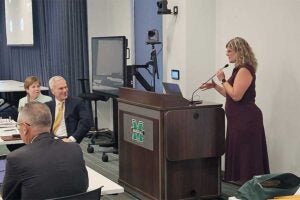 Dr. Rosalynn Quinones speaks at a podium during a Marshall University Board of Governors meeting. Marshall University President Brad Smith and other board members are seated nearby as Quinones presents information about the West Virginia STEM Alliance.