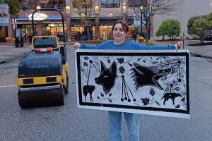 Student holds a large black-and-white relief print featuring coyotes, pawprints, leaves, and tracking graphics, standing beside a pavement roller during the Downtown Huntington Art Walk. The artwork was inspired by Dr. Shane Welch's coyote research.