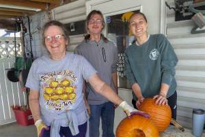 Three Marshall University mathematics volunteers carve pumpkins for the Kenova Pumpkin House display, holding large pumpkins on a worktable.